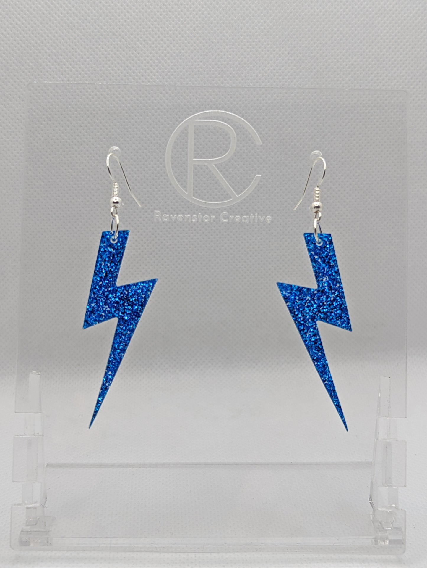 Shows a pair of earrings that have silver ear wire hook fixings. They are Lightning Bolt shapes similar to Harry Potter's forehead. They are Royal Blue Glitter colour and are against a white background on see through stand.