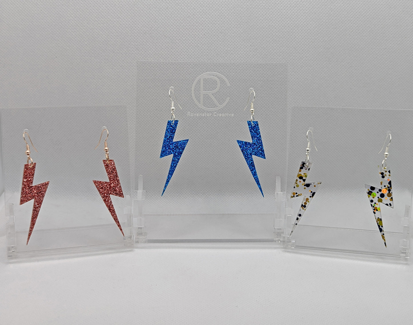 Three pairs of earrings that have ear wire fixings. They are Lightning Bolt shapes similar to Harry Potters forehead. The colours are Copper Glitter, Royal Blue Glitter and Lollipop Glitter. They are against a white background on see through stands.