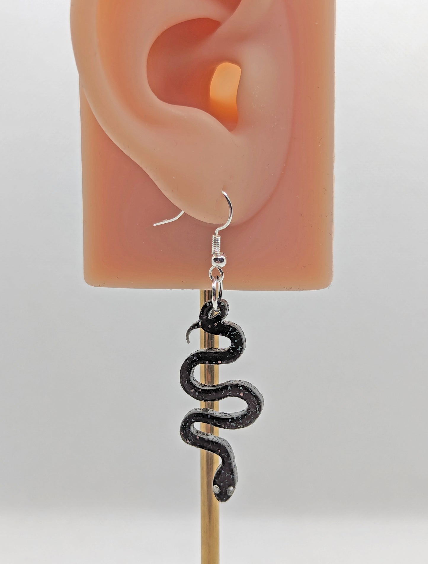 Shows a pair of acrylic earrings with gold ear wire hook fixings. They are hanging snake shapes. The colour is Black & Silver Glitter. They are against an ear to show the size.