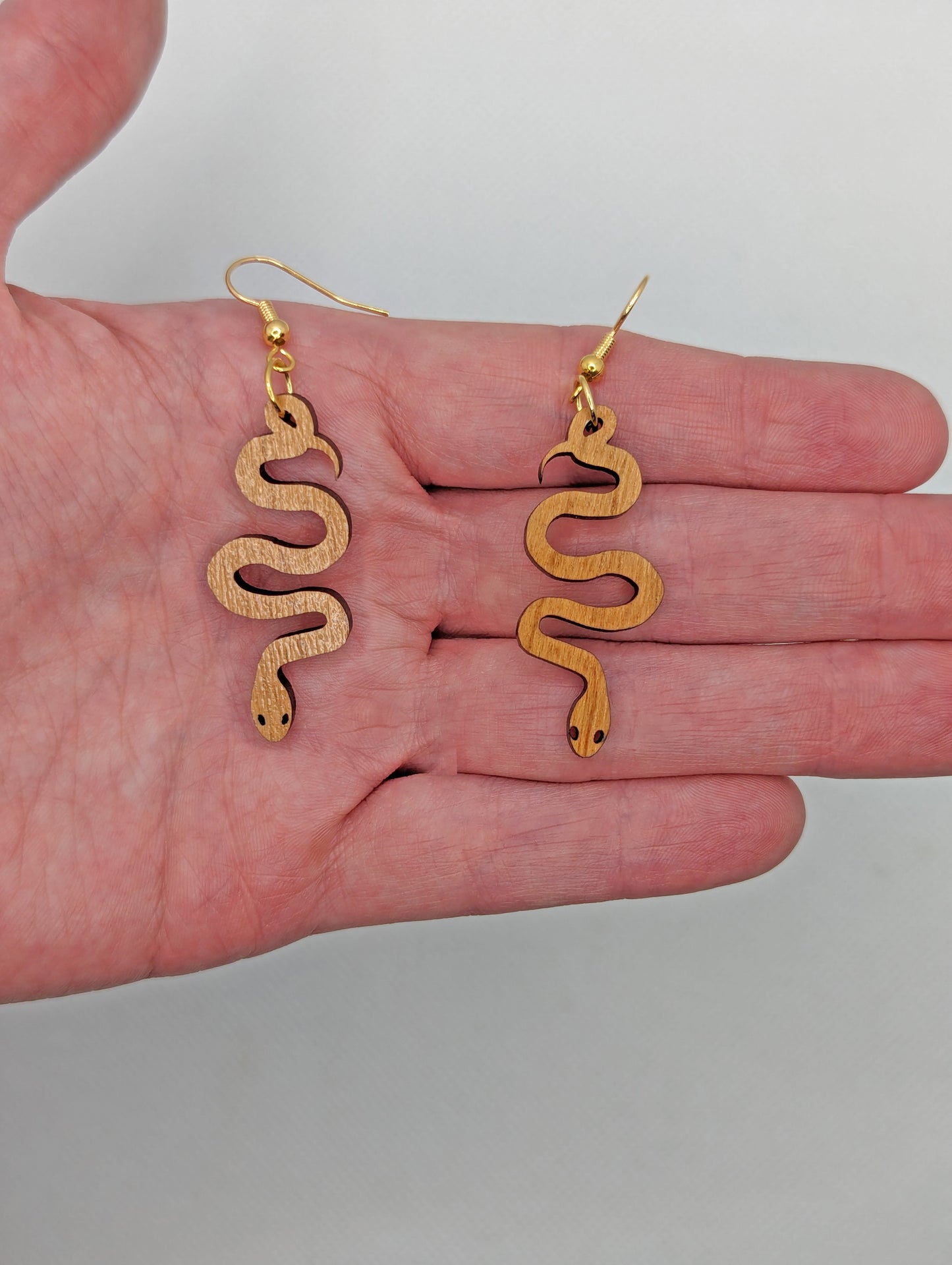 Shows a pair of wooden earrings with gold ear wire hook fixings. They are hanging snake shapes. The colour is Cherry Wood. They are against a hand showing the size of two and a half finger widths in length and one wide.