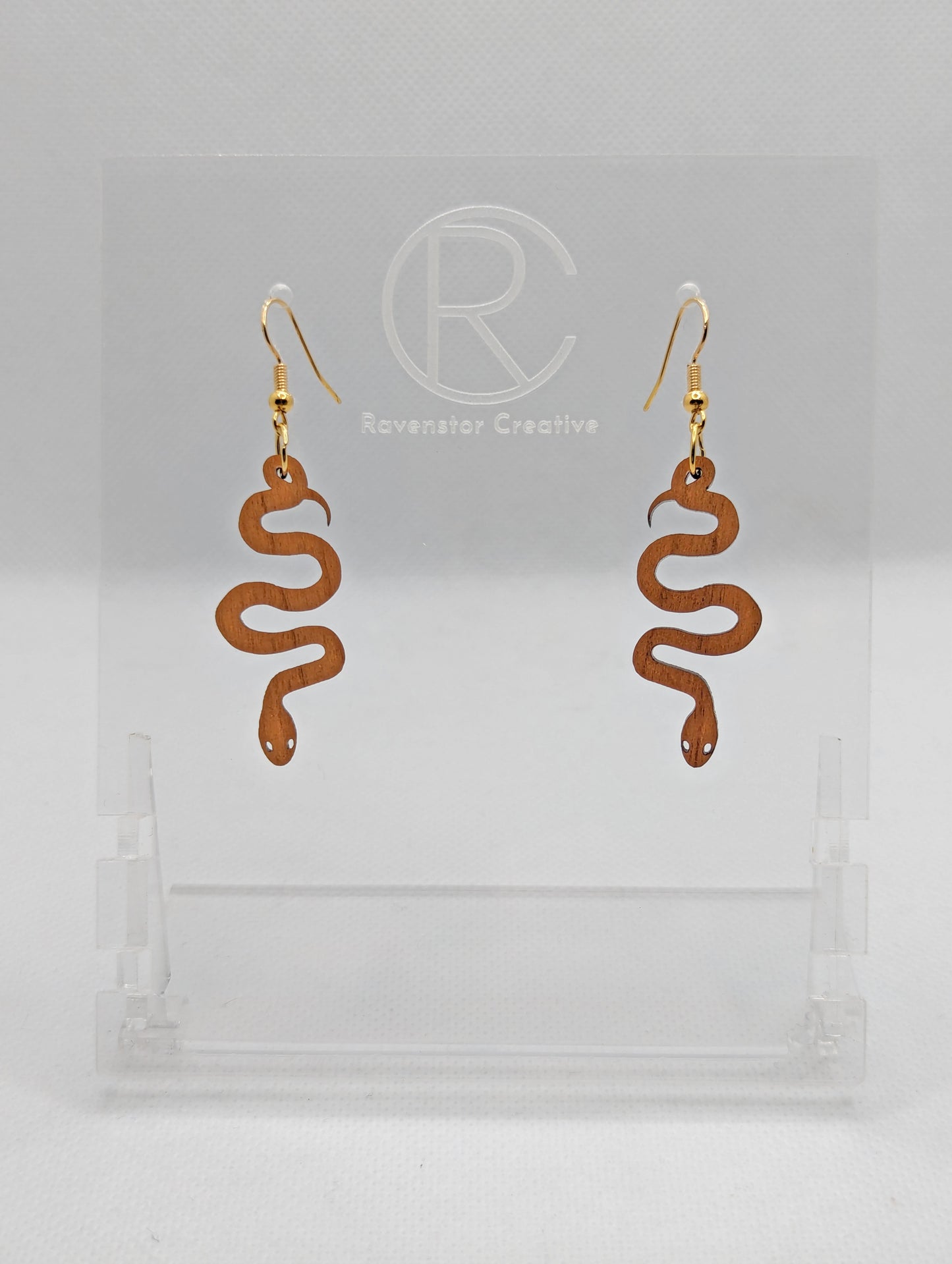 Shows a pair of wooden earrings with gold ear wire hook fixings. They are hanging snake shapes. The colour is Cherry Wood. They are against a white background on a clear stand with the Ravenstor Creative logo on it.