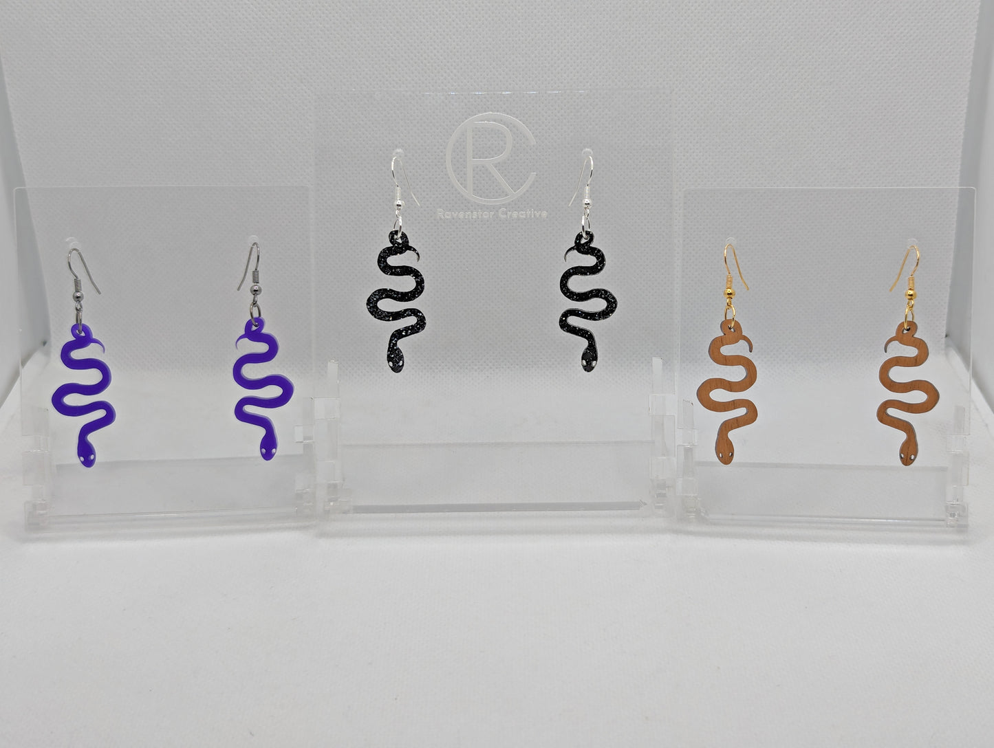 Shows three pairs of earrings with ear wire hook fixings. They are hanging snake shapes. The colours are Purple Acrylic, Black & Silver Glitter Acrylic and Cherry Wood. They are against a white background on a clear stand.