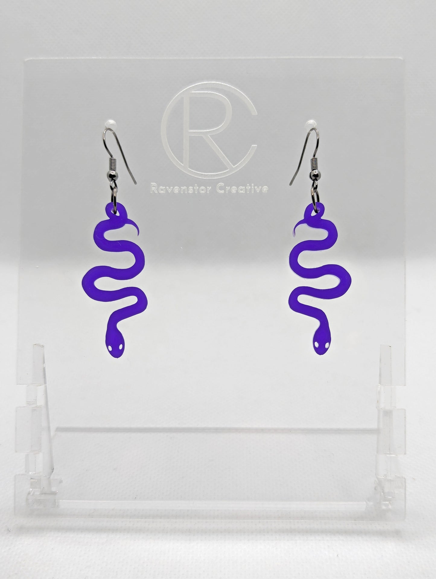 Shows a pair of Acrylic earrings with black ear wire hook fixings. They are hanging snake shapes. The colour is purple. They are against a white background on a clear stand with the Ravenstor Creative logo on it.