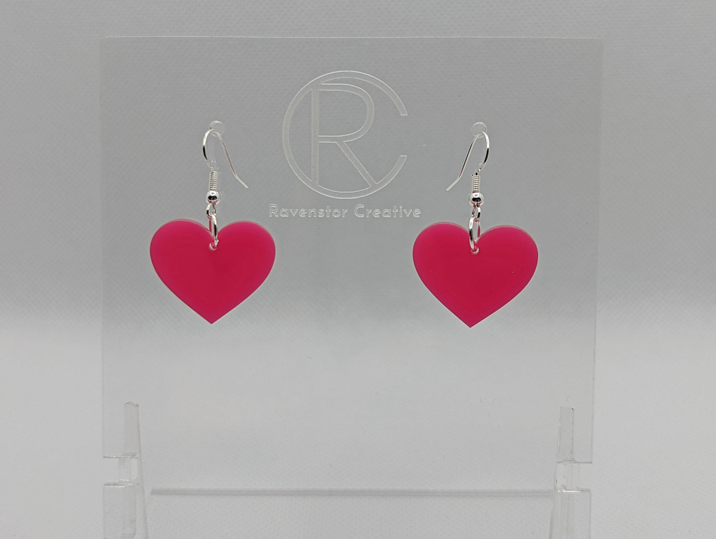 Shows a pair of acrylic earrings with silver ear wire hook fixings. They are a heart shape. The colour of them is pretty hot pink. They are on a see through stand that has the Ravenstor Creative Logo and its against a white background.