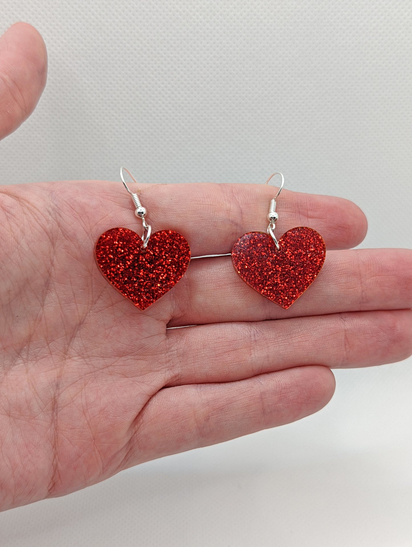 Image of a pair of heart shaped earrings on a hand showing the size against a white background, they are one and a half finger widths length and the same again wide. They are Red glitter and have silver fixings.