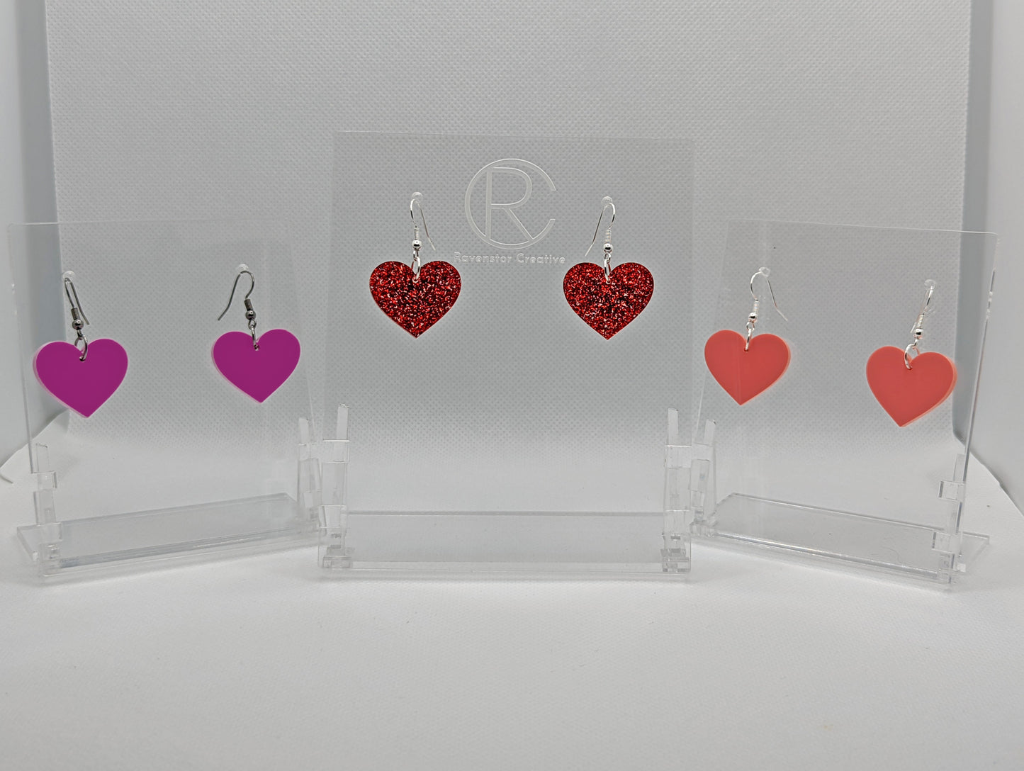 Shows three pairs of earrings that have ear wire hook fixings. They are pretty love Heart shaped. The colours are Grape, red glitter and sherbet pink. They are against a white background on see through stand.