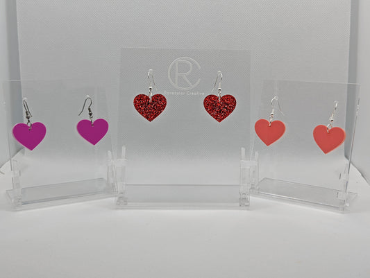 Shows three pairs of earrings that have ear wire hook fixings. They are pretty love Heart shaped. The colours are Grape, red glitter and sherbet pink. They are against a white background on see through stand.