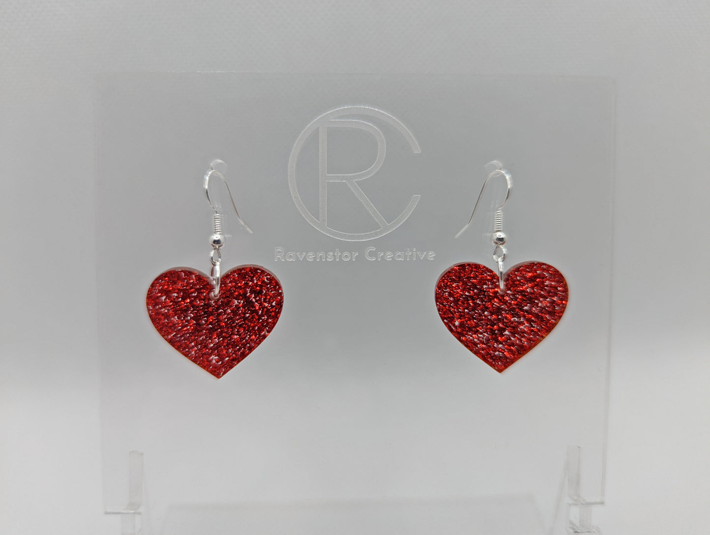 Shows a pair of acrylic earrings with silver ear wire hook fixings. They are a heart shape. The colour of them is sparkly red glitter. They are on a see through stand that has the Ravenstor Creative Logo and its against a white background.