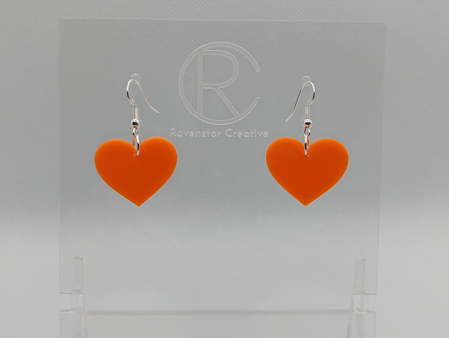 Shows a pair of acrylic earrings with silver ear wire hook fixings. They are a heart shape. The colour of them is a striking orange. They are on a see through stand that has the Ravenstor Creative Logo and its against a white background.
