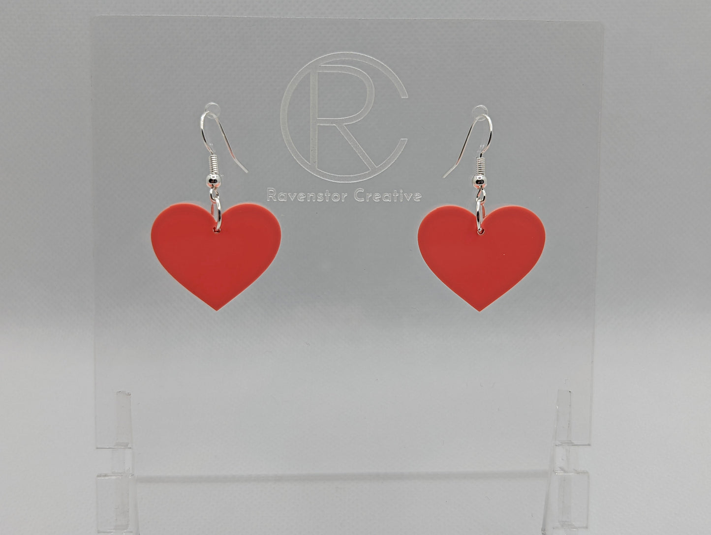 Shows a pair of acrylic earrings with silver ear wire hook fixings. They are a heart shape. The colour of them is pretty sherbet light pink. They are on a see through stand that has the Ravenstor Creative Logo and its against a white background.