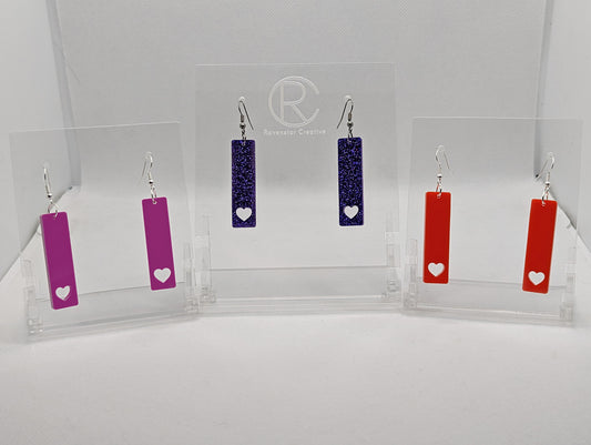 Shows three pairs of earrings that have ear wire fixings. They are slim long bar pendant earrings with a heart at the bottom. The colours are grape, purple glitter and red. They are against a white background on see through stand.