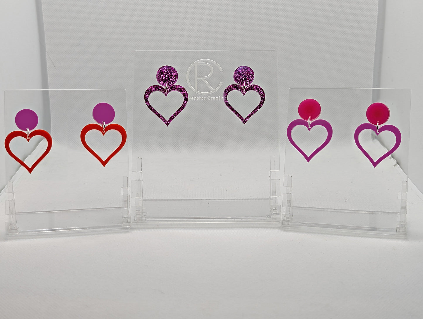 Shows three pair earrings with stud fixings attached to an acrylic circle, dangling under is a coloured heart, cut-out in the middle. The colours are grape Circle with orange heart, Pink glitter circle/matching heart and a hot pink circle/grape heart