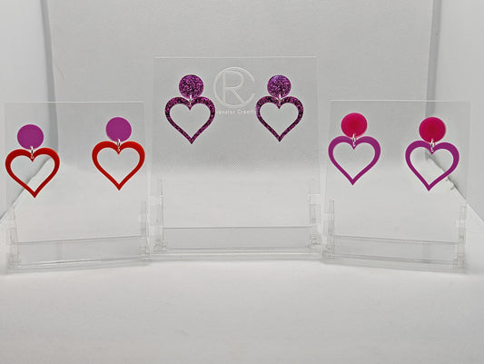 Shows three pair earrings with stud fixings attached to an acrylic circle, dangling under is a coloured heart, cut-out in the middle. The colours are grape Circle with orange heart, Pink glitter circle/matching heart and a hot pink circle/grape heart