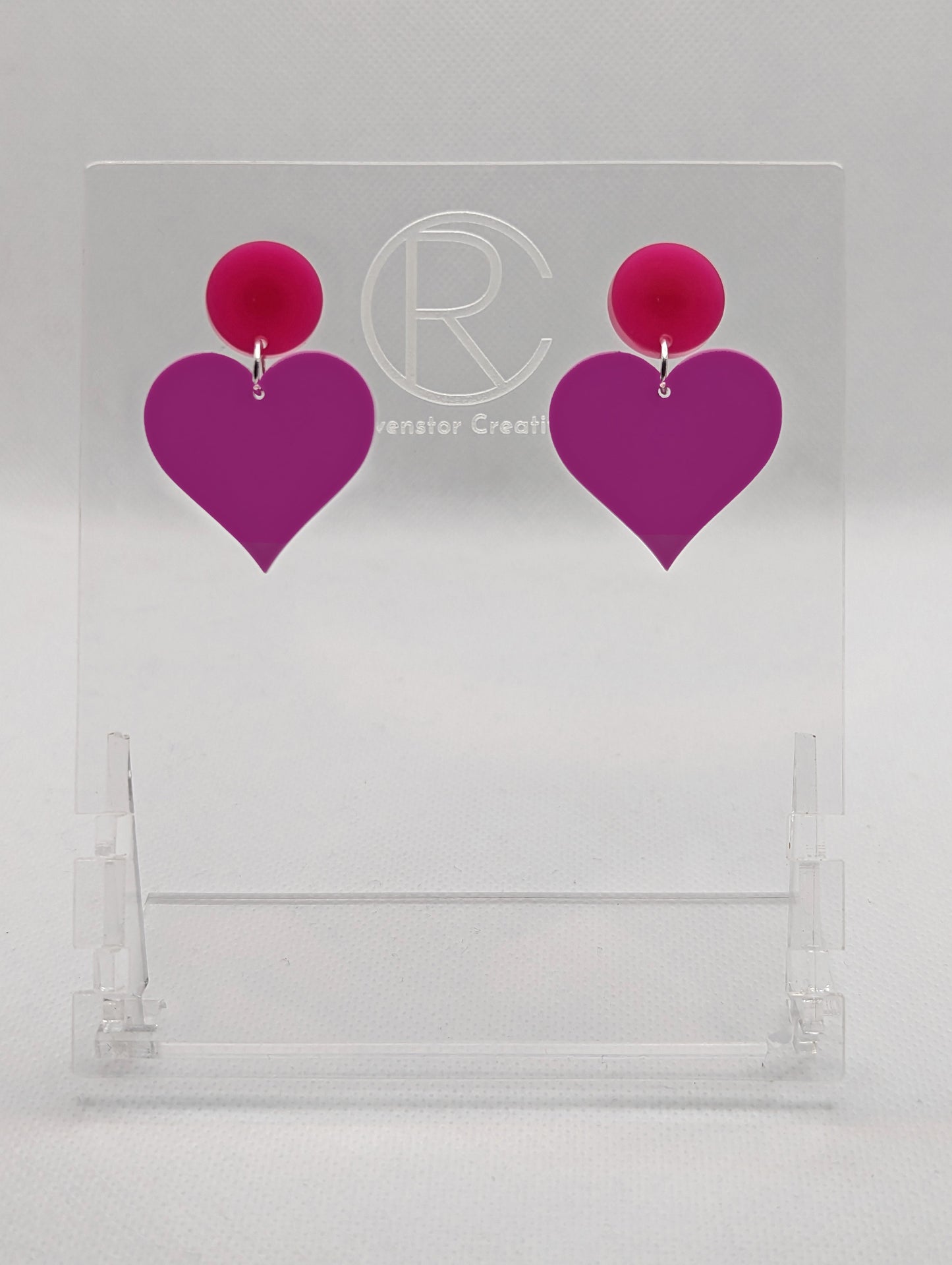 Shows a pair of earrings that are two different colours with stud fixings attached to an acrylic circle, dangling under is a coloured heart. The colours are a hot pink circle and grape heart dangling underneath