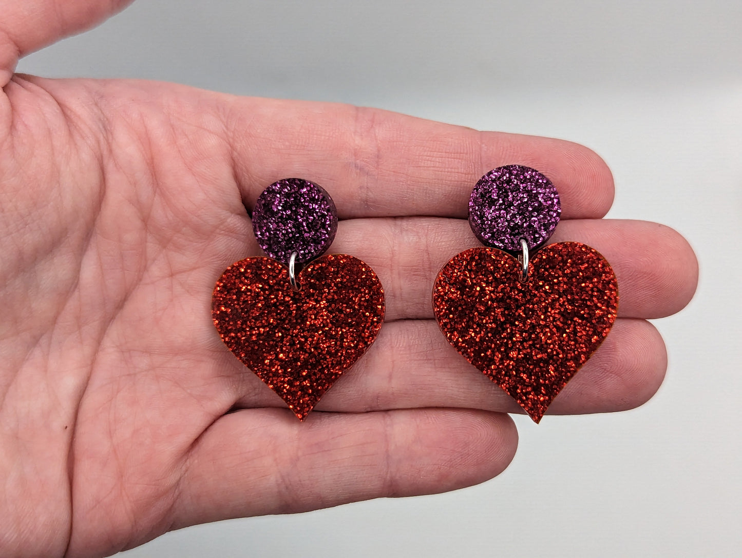 Earrings with stud fixings on a hand to show the size. They are 3 finger widths length and 2 wide. They are an acrylic circle, with a heart dangling underneath. The colour is a Glitter Pink circle with red glitter heart dangling underneath.