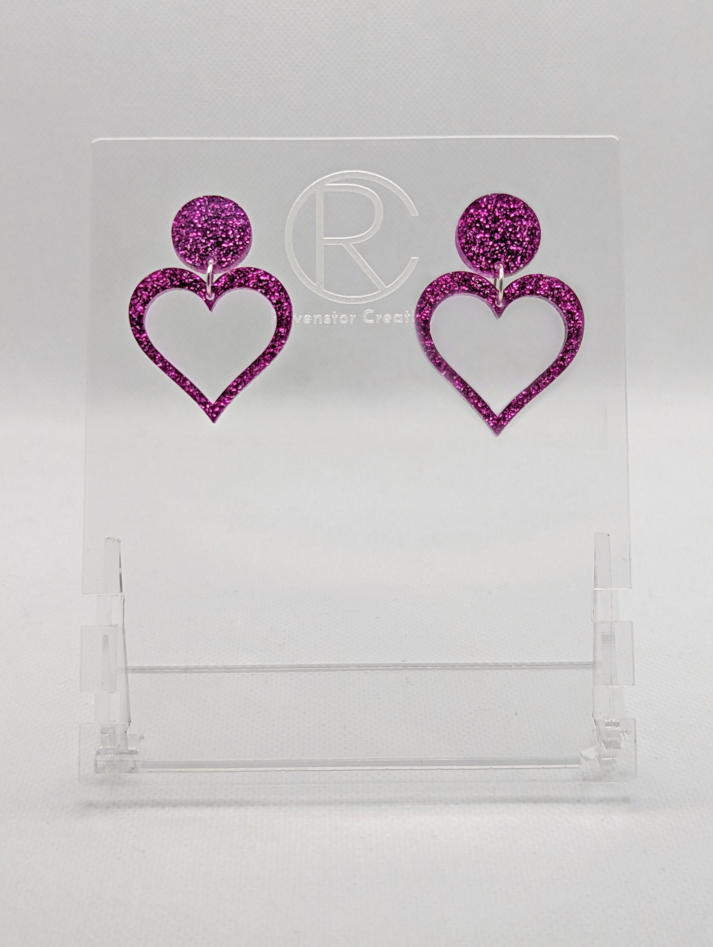 Shows earrings with stud fixings attached to an acrylic circle, dangling under is a coloured heart cut-out in the middle. The colour is a Glitter Pink circle with matching heart dangling underneath.