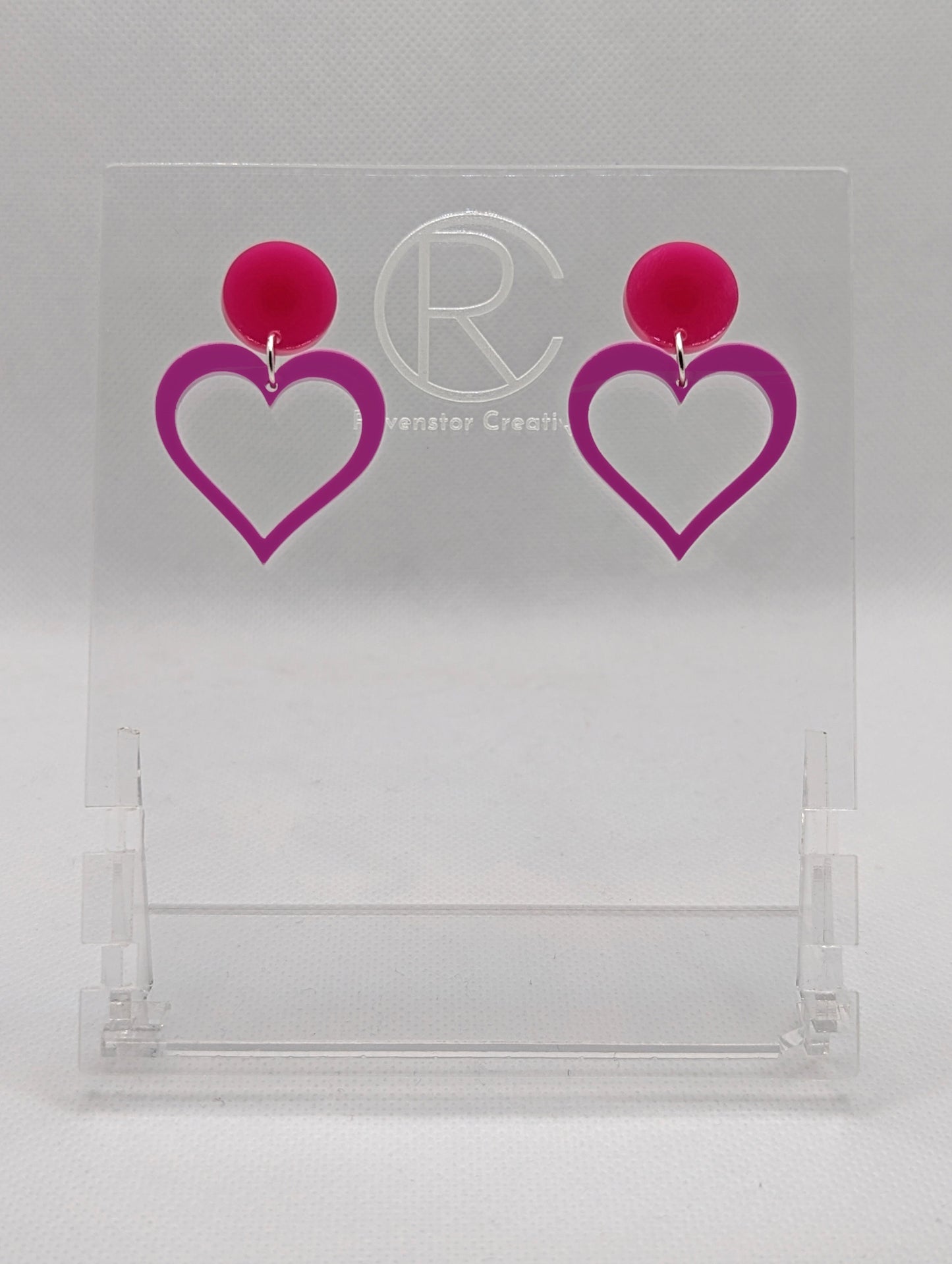 Shows earrings with stud fixings attached to an acrylic circle, dangling under is a coloured heart cut-out in the middle. The colour is a hot pink circle with a grape heart dangling underneath.