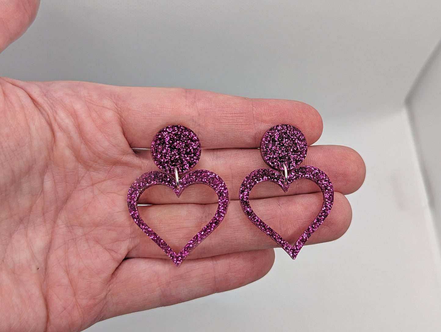 Earrings with stud fixings on a hand to show the size. They are 3 finger widths length and 2 wide. They are an acrylic circle, with a heart that's cut-out in the middle. The colour is a Glitter Pink circle with matching heart dangling underneath.
