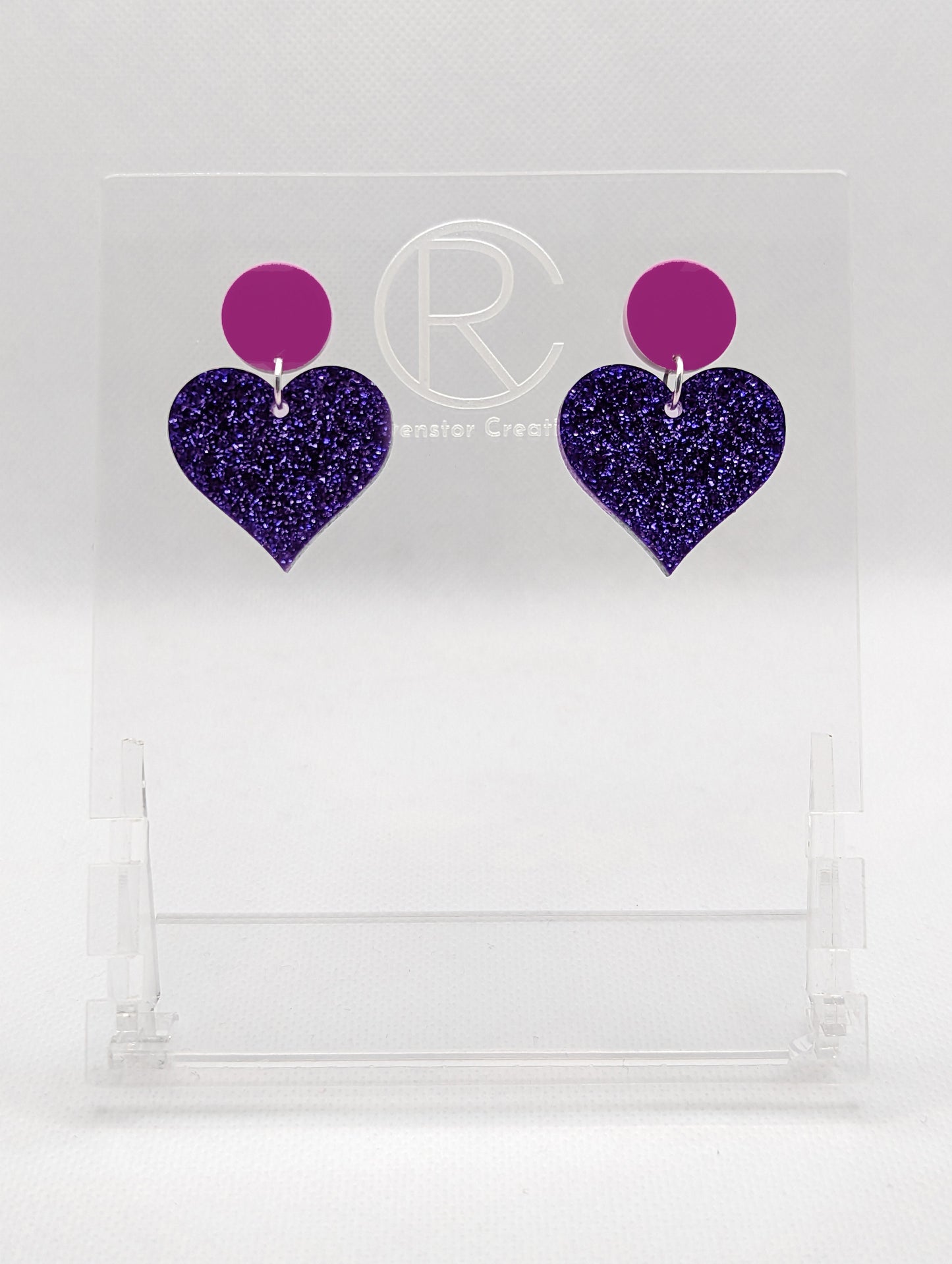 Shows a pair of earrings that are two different colours with stud fixings attached to an acrylic circle, dangling under is a coloured heart. The colours are a grape circle and purple glitter heart dangling underneath