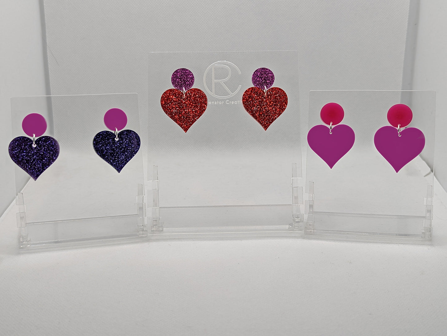 Shows three pairs earrings with stud fixings attached to an acrylic circle, dangling under is a coloured heart. The colours are grape Circle with purple glitter heart, Pink glitter circle and red glitter heart and a hot pink circle and grape heart