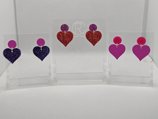 Shows three pairs earrings with stud fixings attached to an acrylic circle, dangling under is a coloured heart. The colours are grape Circle with purple glitter heart, Pink glitter circle and red glitter heart and a hot pink circle and grape heart