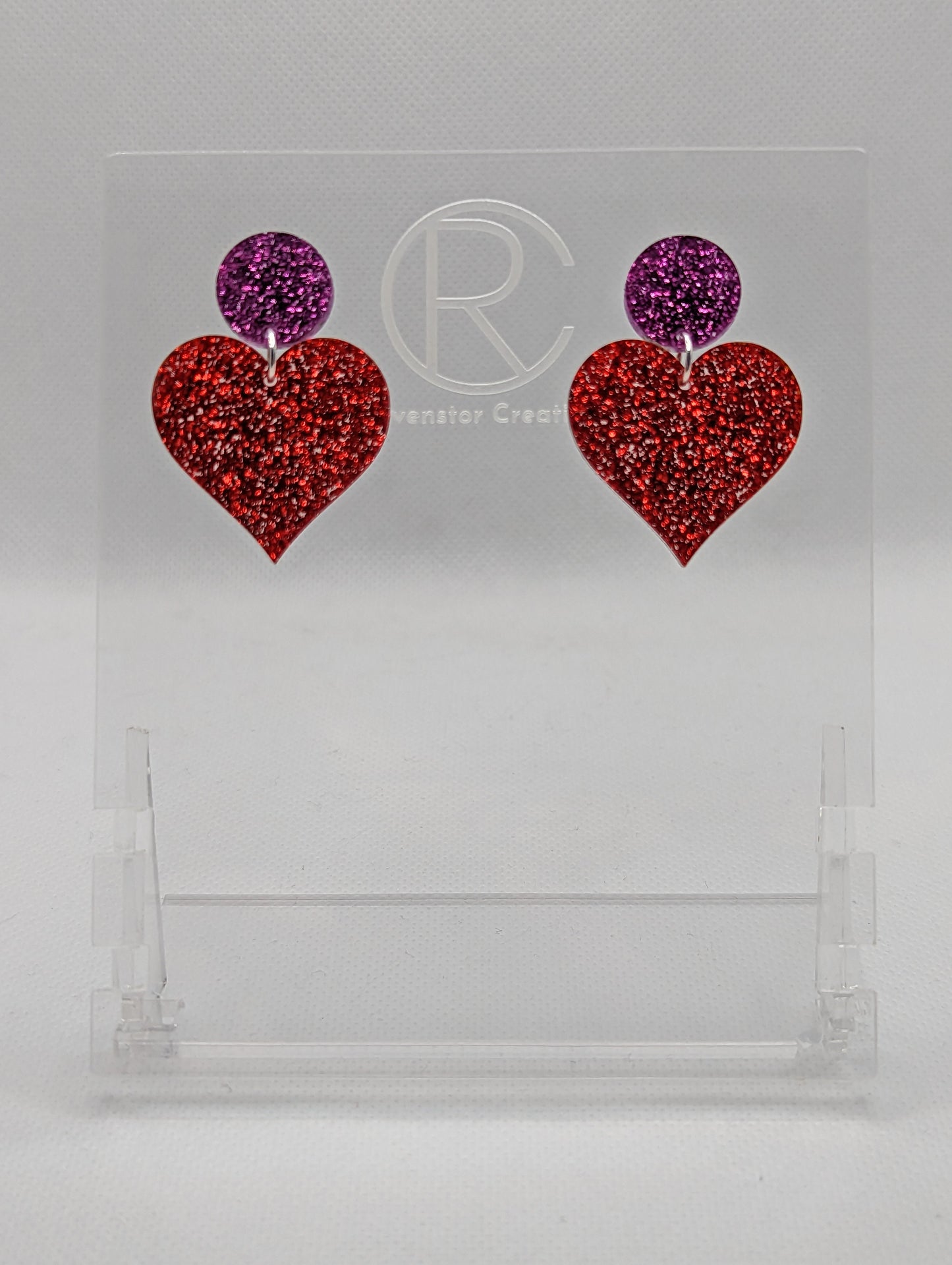 Shows a pair of earrings that are two different colours with stud fixings attached to an acrylic circle, dangling under is a coloured heart. The colours are a pink glitter circle and red glitter heart dangling underneath