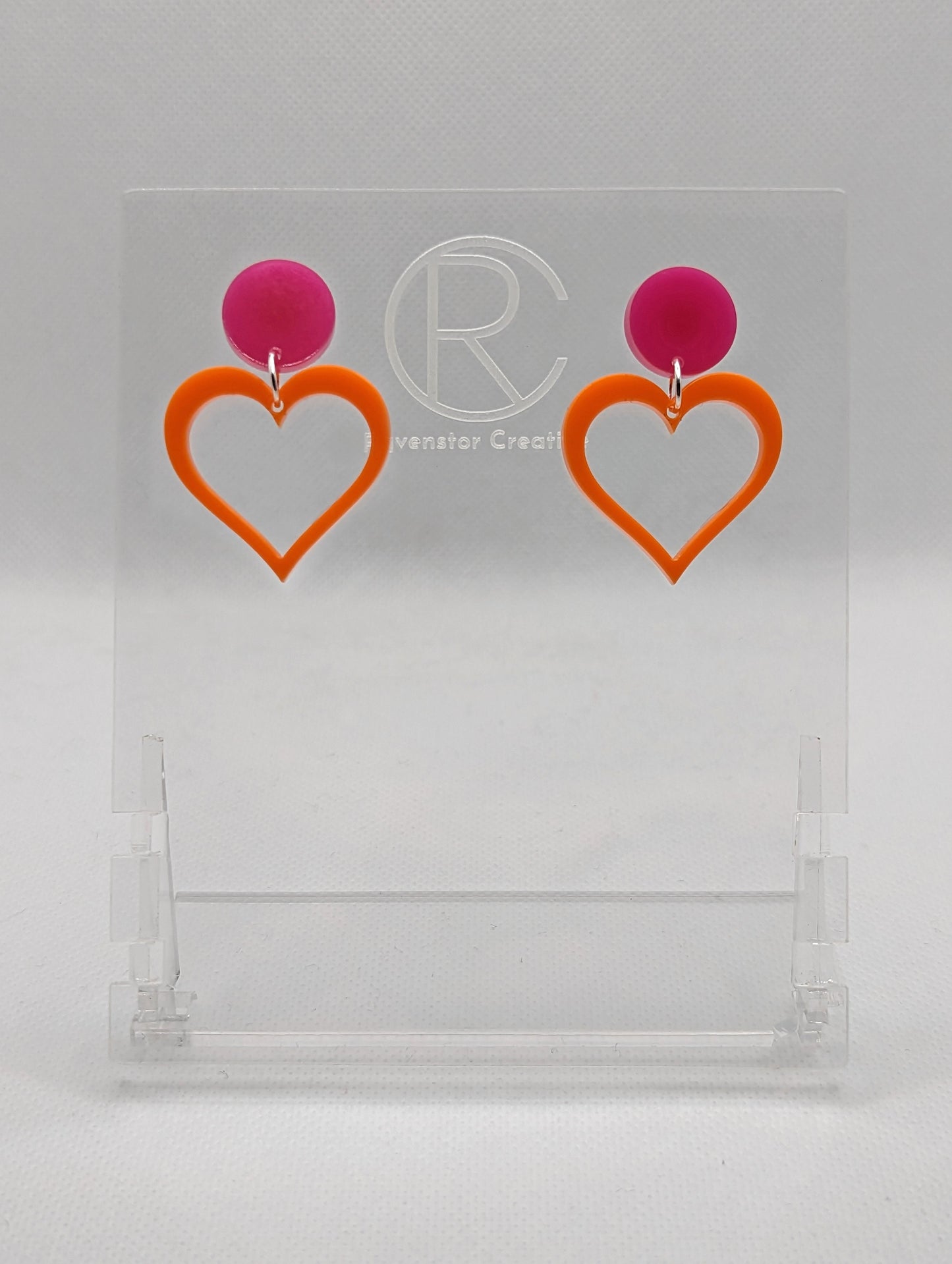 Shows earrings with stud fixings attached to an acrylic circle, dangling under is a coloured heart cut-out in the middle. The colour is a hot pink circle with an orange heart dangling underneath.