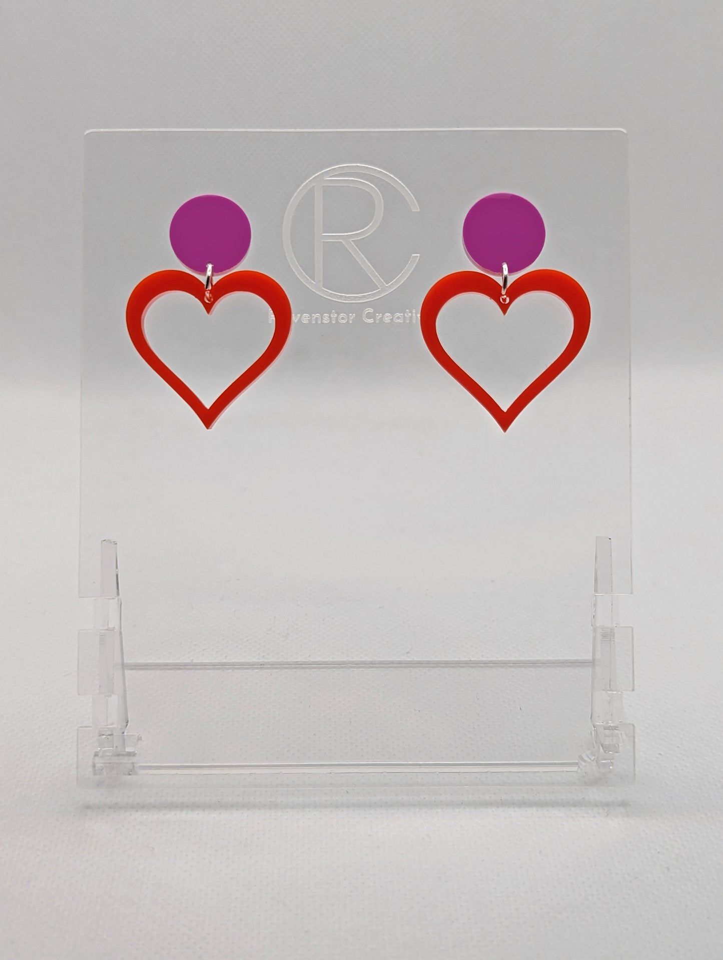 Shows earrings with stud fixings attached to an acrylic circle, dangling under is a coloured heart cut-out in the middle. The colour is a grape circle with a red heart dangling underneath.
