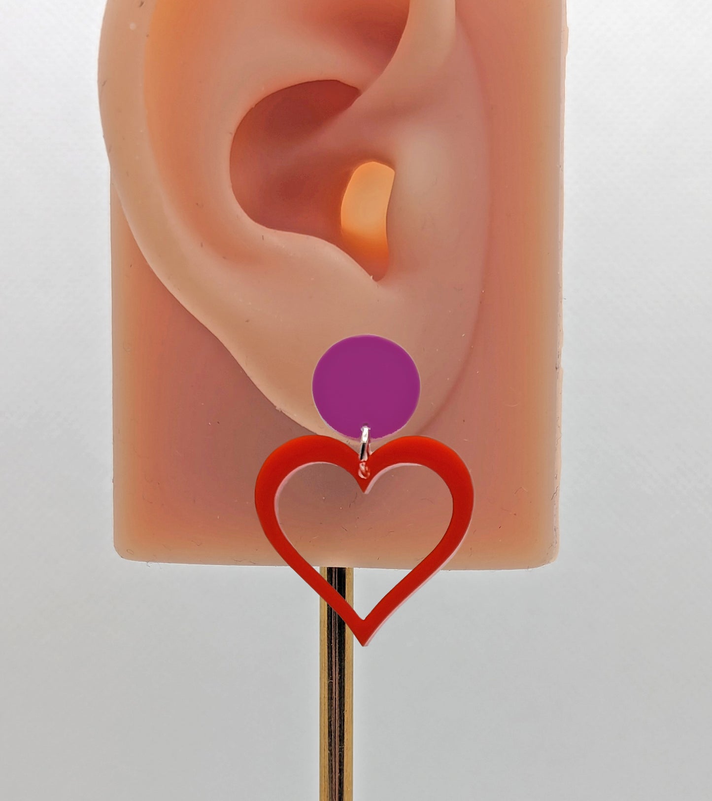 An earrings with stud fixings on an ear to show the size. It is an acrylic circle, with a heart that is cut-out in the middle. The colour is a grape circle with a red heart dangling underneath.