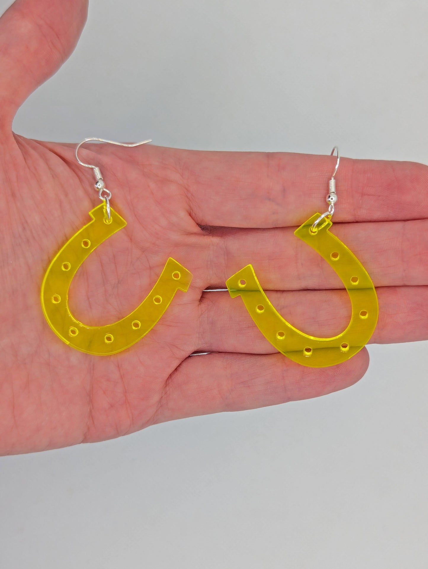 Shows a pair of acrylic earrings that have silver ear wire hook fixings. They are lucky horseshoe St Patricks day themed earrings. They are Neon Orange in colour and are against a hand to show the size of two and a half finger widths in length and width.