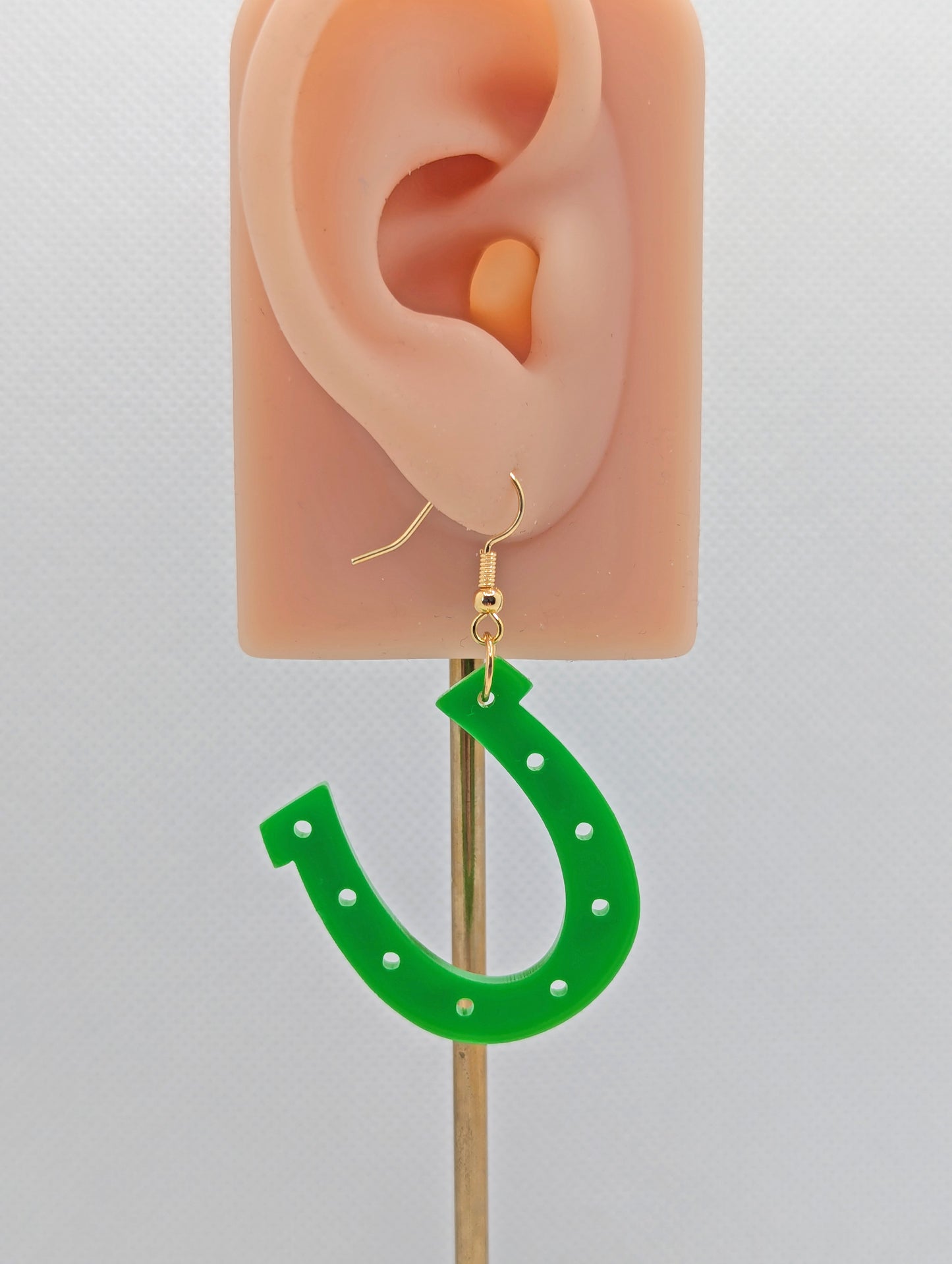 Image of a lucky horseshoe St Patricks day themed earring on an ear showing the size against a white background. They are made from Green acrylic and have gold ear wire hook fixings.
