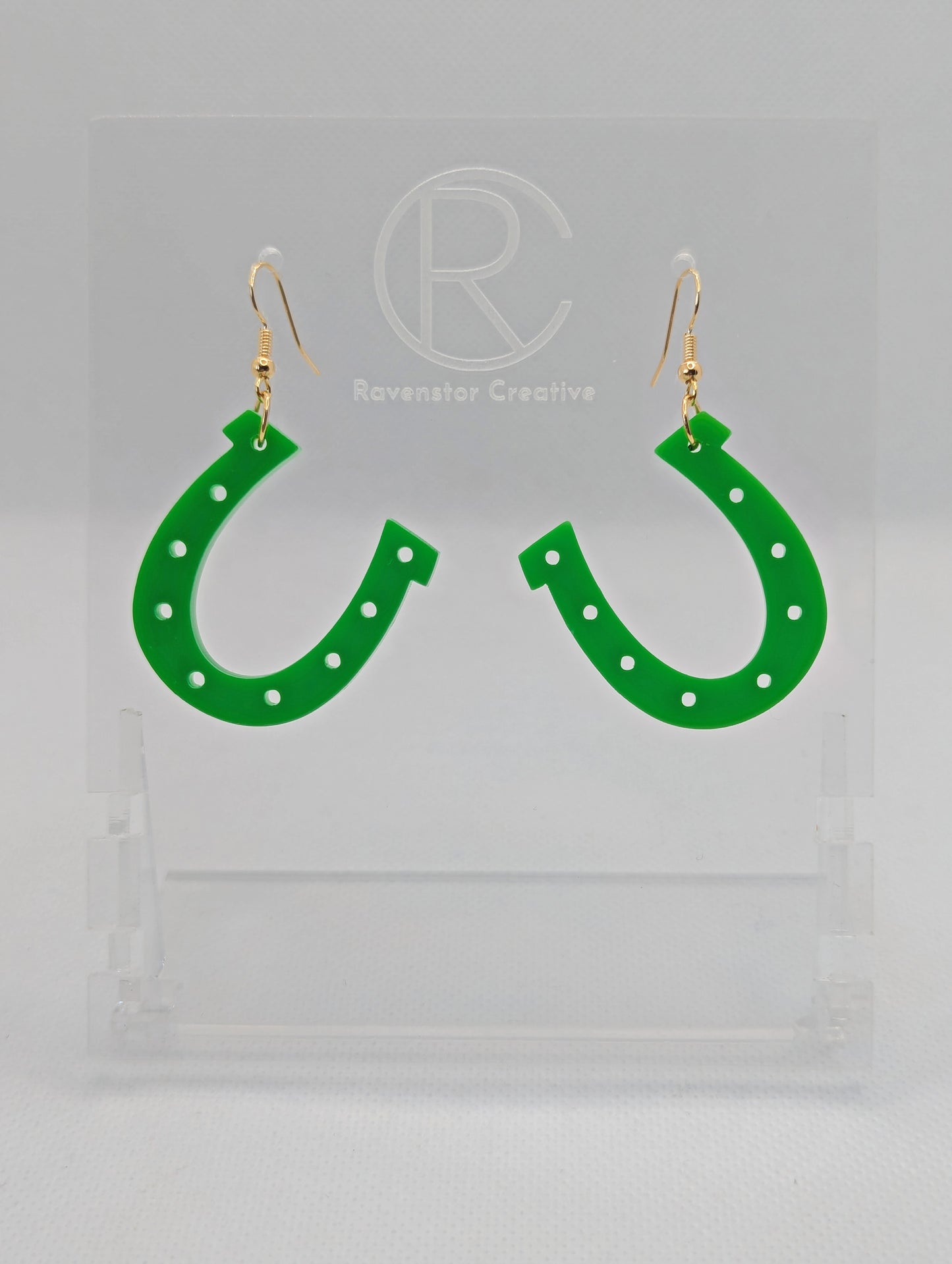 Shows a pair of acrylic earrings that have gold ear wire hook fixings. They are lucky horseshoe St Patricks day themed earrings. They are Green in colour and are against a white background on a see through stand.