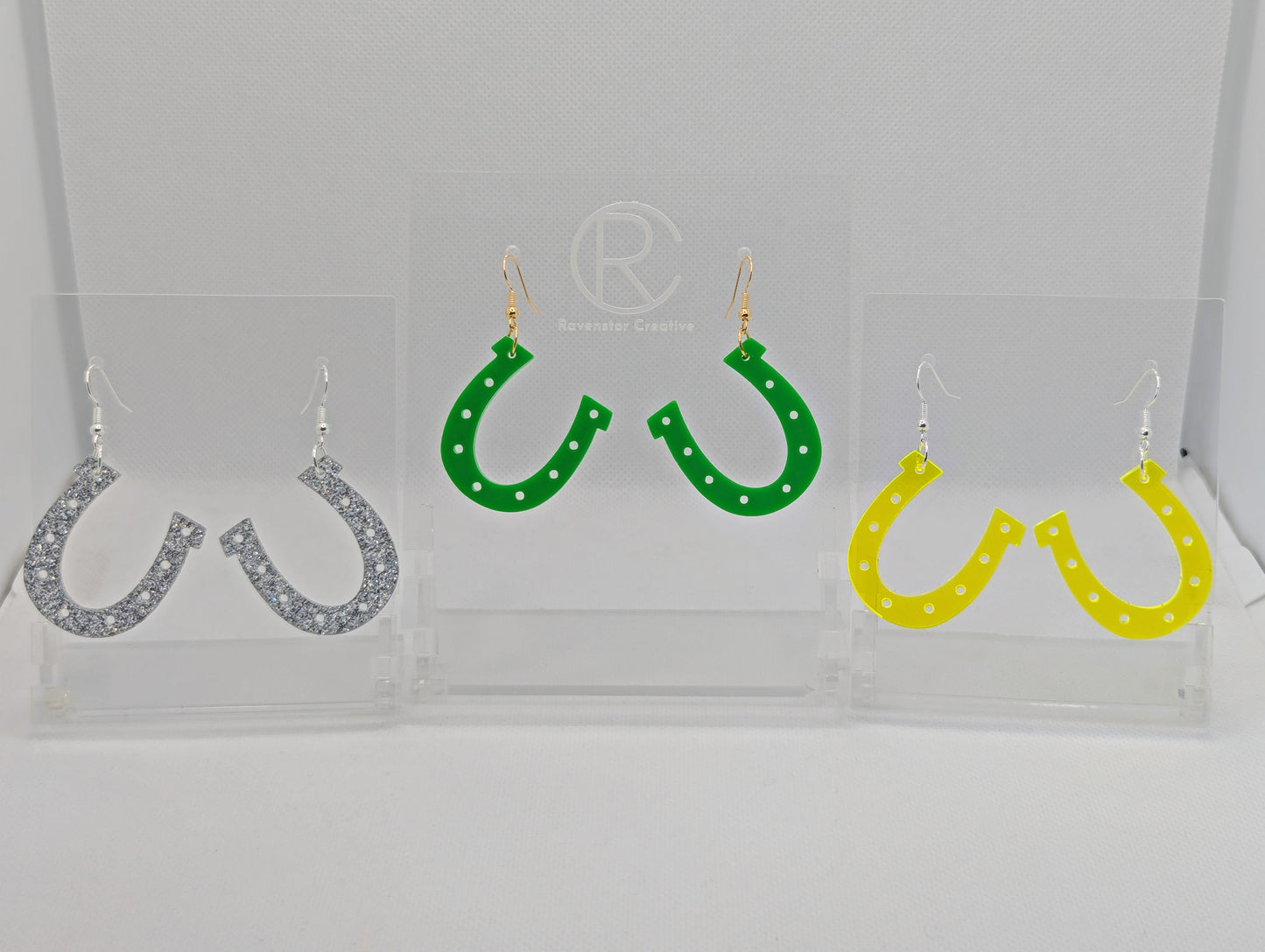 Shows three pairs of earrings that have ear wire hook fixings. They are lucky horseshoe earrings. The colours are neon orange, Green and silver glitter. They are against a white background on see through stands.