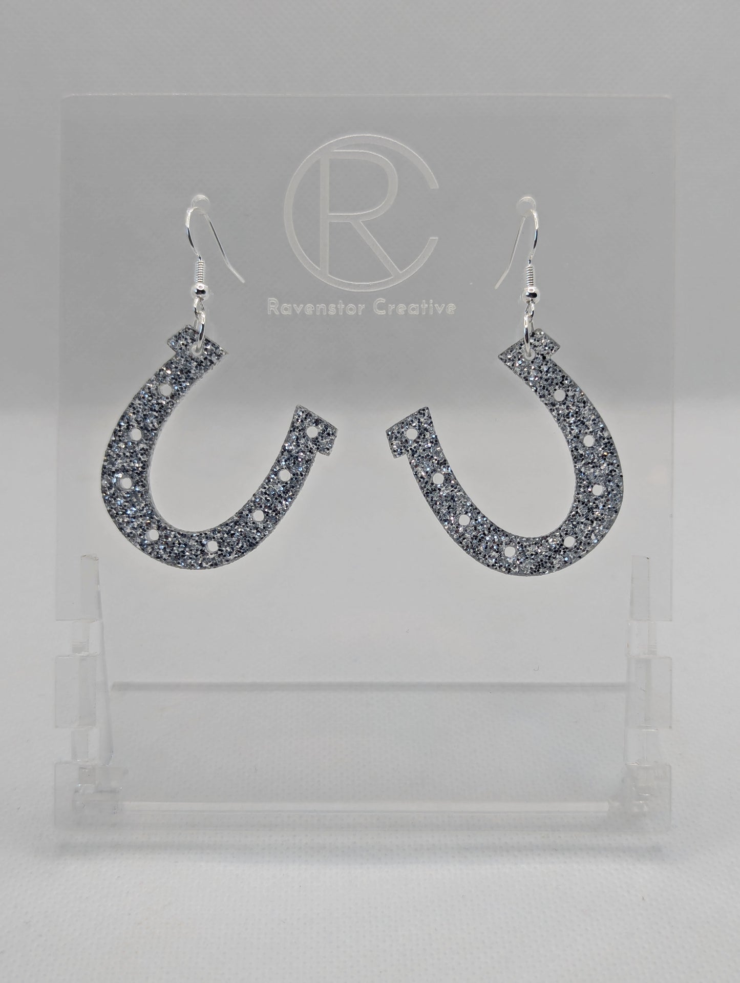 Shows a pair of acrylic earrings that have silver ear wire hook fixings. They are lucky horseshoe St Patricks day themed earrings. They are Silver Glitter in colour and are against a white background on a see through stand.