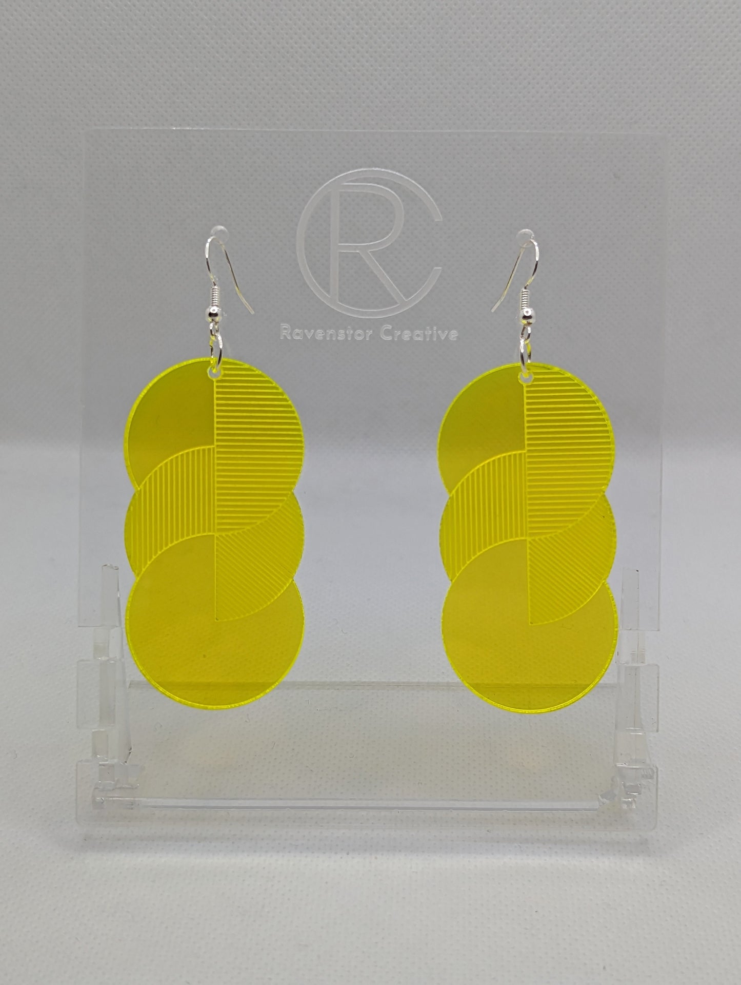 A pair of large acrylic earrings with silver ear wire hook fixings. Each earring is 3 circles overlapping and entwined with engraved line decorations. The colour is neon yellow and they are hung on a clear stand against a white background.