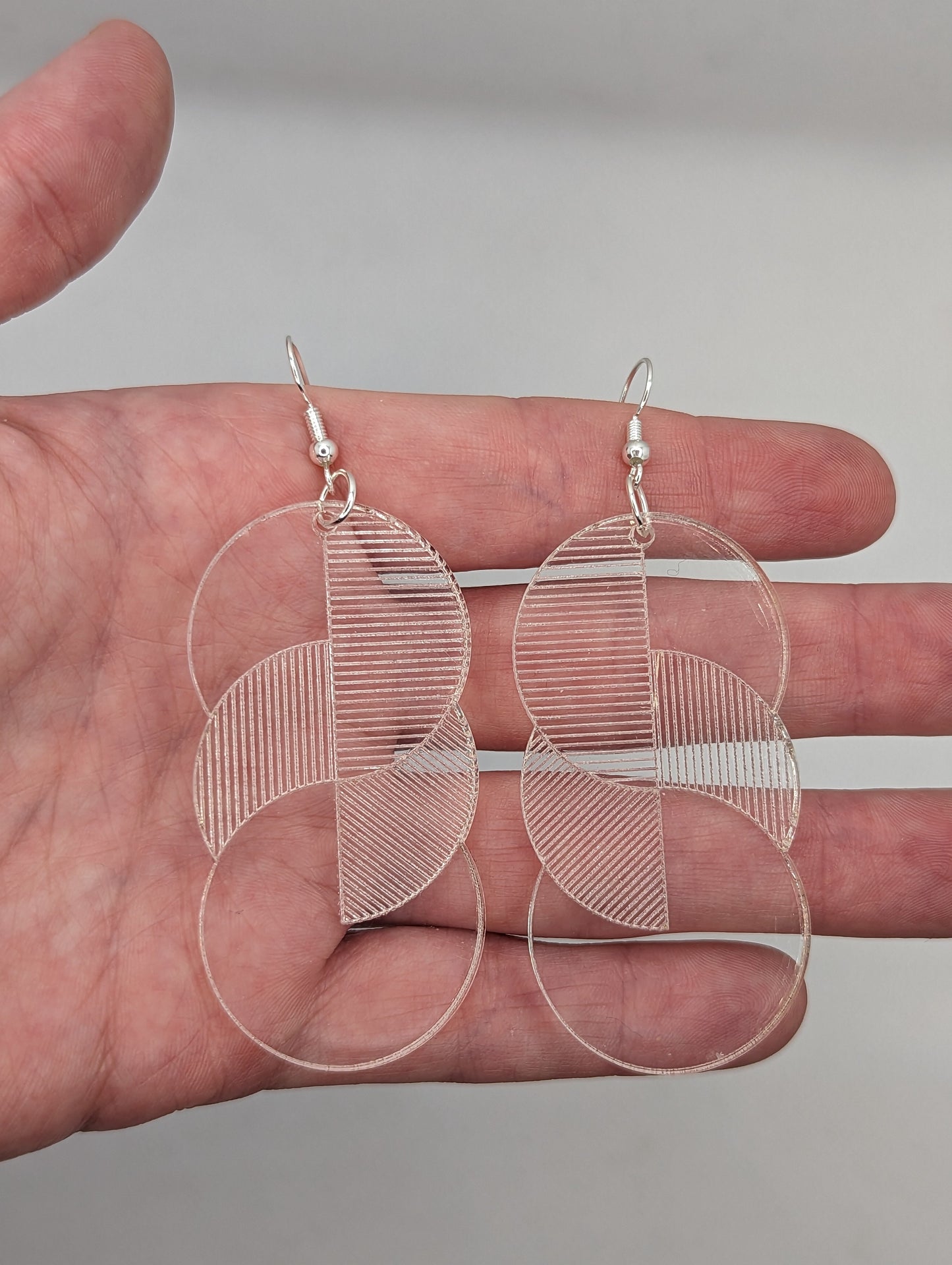 A pair of large clear acrylic earrings with silver ear wire hook fixings against a hand to show the size. Each one is 3 circles overlapping and entwined with engraved line decorations. They are four finger widths in length and two wide.