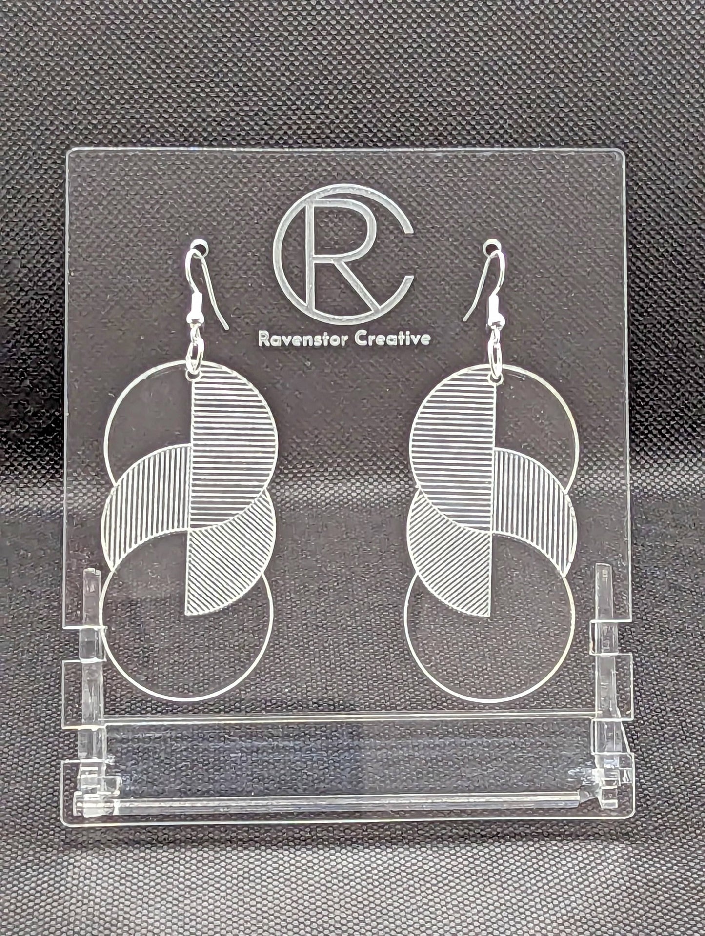 A pair of large acrylic earrings with silver ear wire hook fixings. Each earring is 3 circles overlapping and entwined with engraved line decorations. The colour is clear / See through and they are hung on a clear stand against a black background.