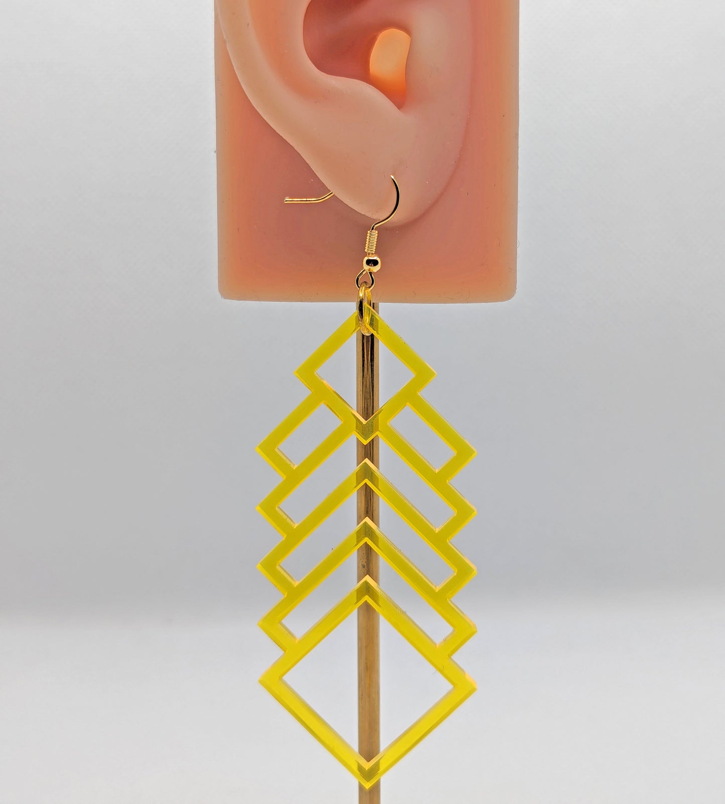 A  large and long acrylic geometric diamond themed earring. It is neon orange in colour and has gold ear wire hook fixings and is on an ear to show the size against a white background.