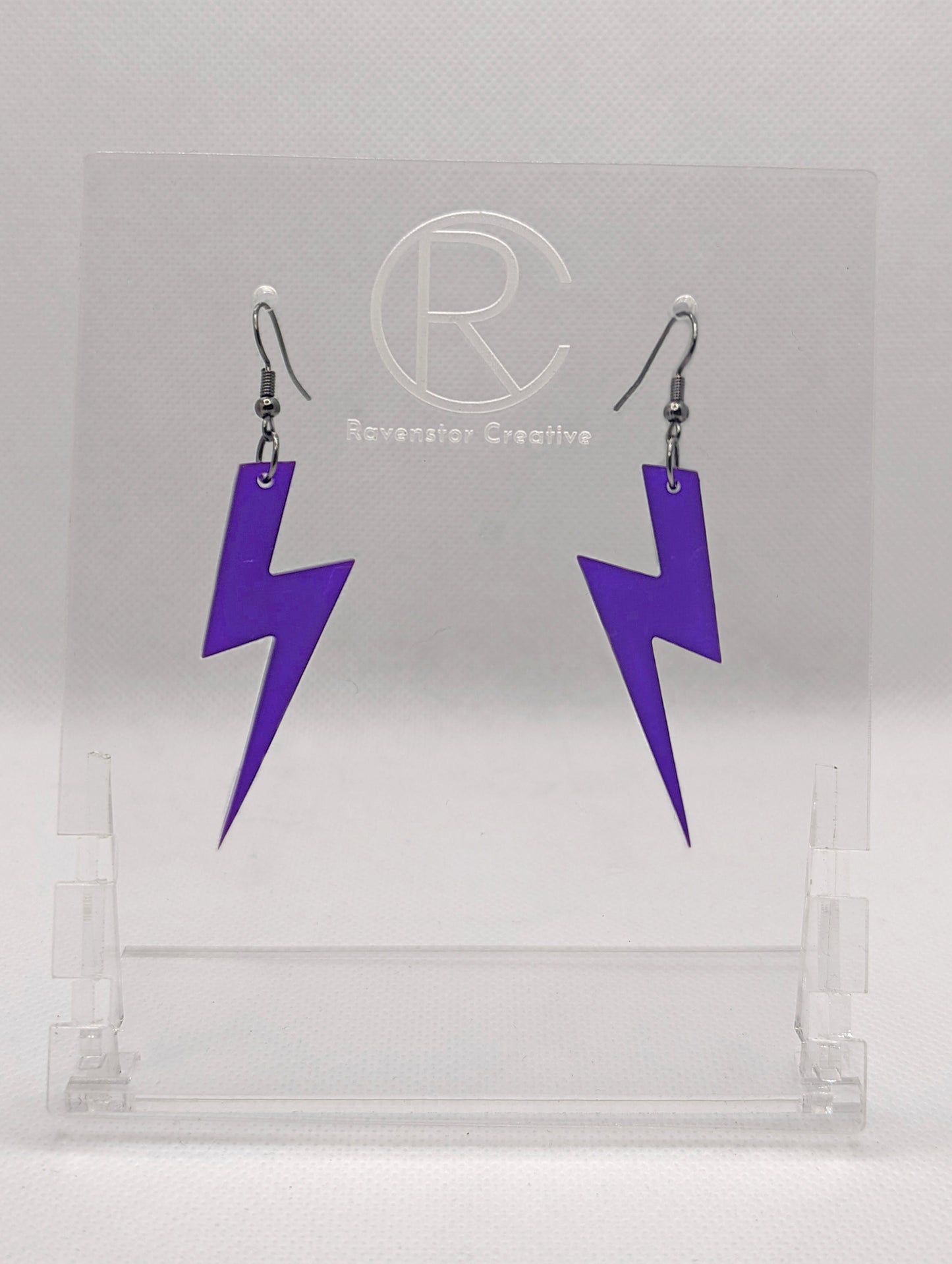 Shows a pair of acrylic earrings that have silver ear wire hook fixings. They are Lightning Bolt shapes similar to Harry Potter's forehead. They are purple colour and are against a white background on see through stand.