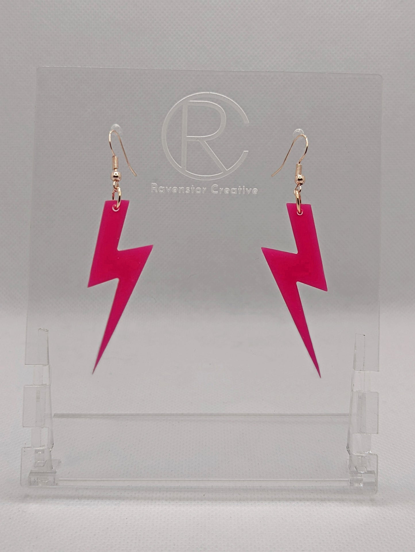 Shows a pair of acrylic earrings that have silver ear wire hook fixings. They are Lightning Bolt shapes similar to Harry Potter's forehead. They are hot pink colour and are against a white background on see through stand.