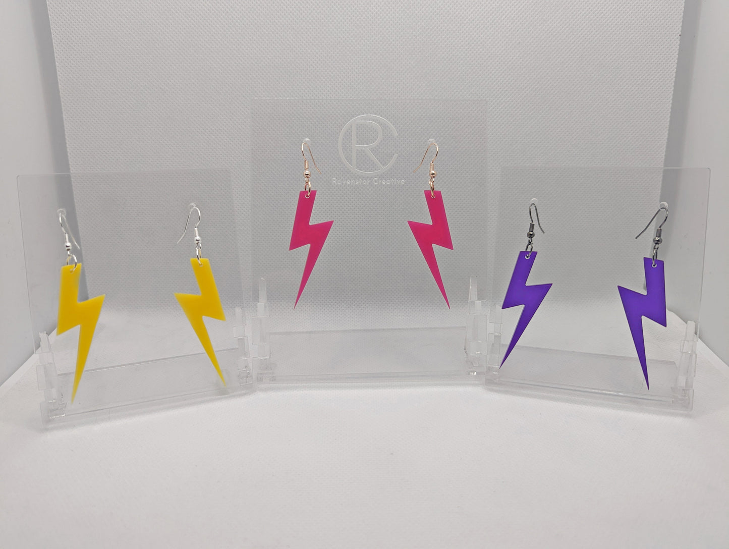 Shows three pairs of acrylic earrings that have ear wire hook fixings. They are Lightning Bolt shapes similar to Harry Potter's forehead. The colours are yellow, hot pink and purple. They are against a white background on see through stands.