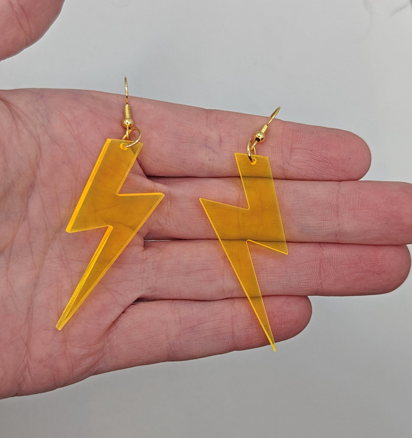 Image of a pair of acrylic lightning bolt shaped earrings on a hand showing the size against a white background, they are three and a half finger widths length and one and a half wide. They are neon orange and have gold fixings.