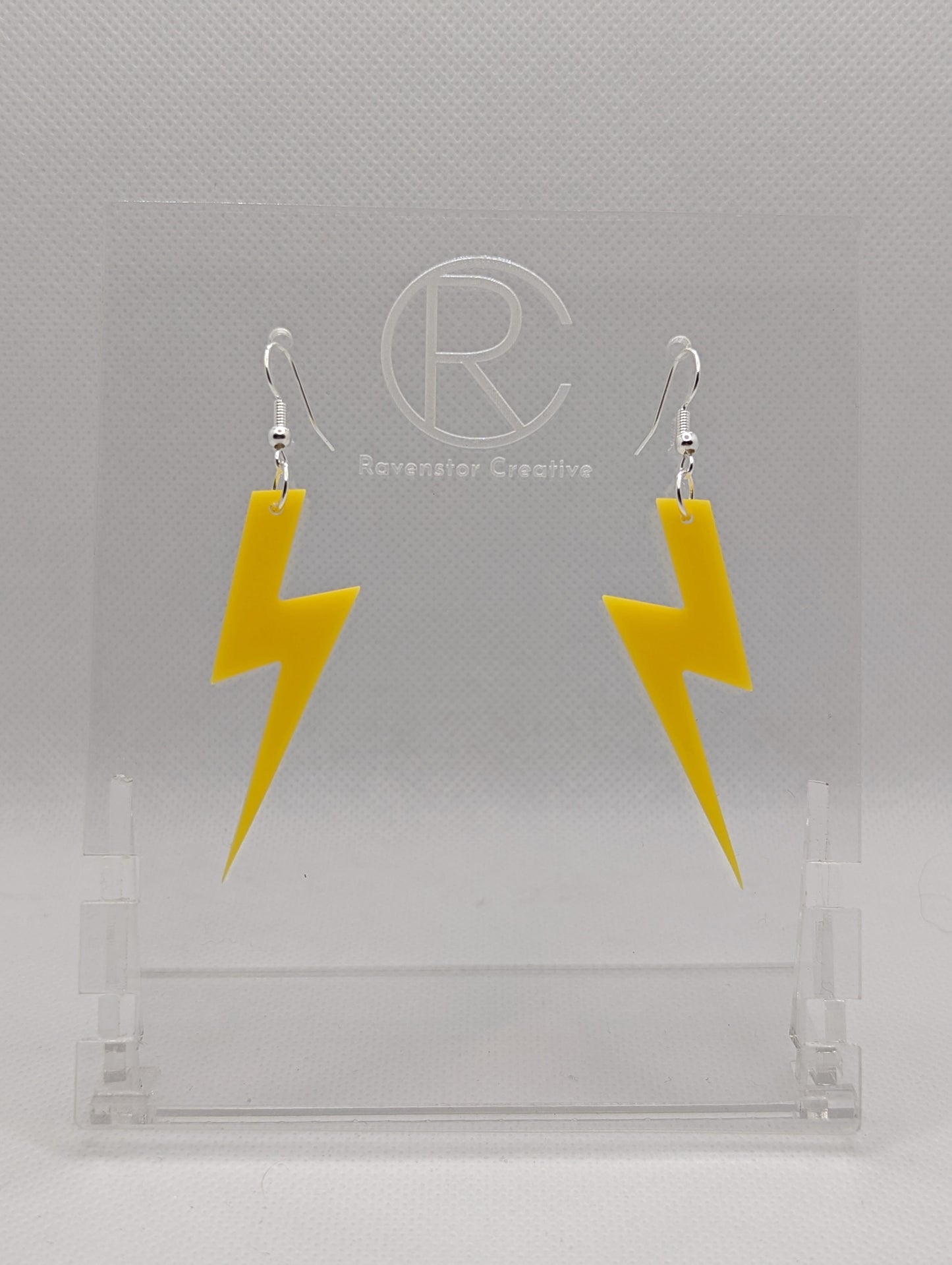 Shows a pair of acrylic earrings that have silver ear wire hook fixings. They are Lightning Bolt shapes similar to Harry Potter's forehead. They are yellow colour and are against a white background on see through stand.