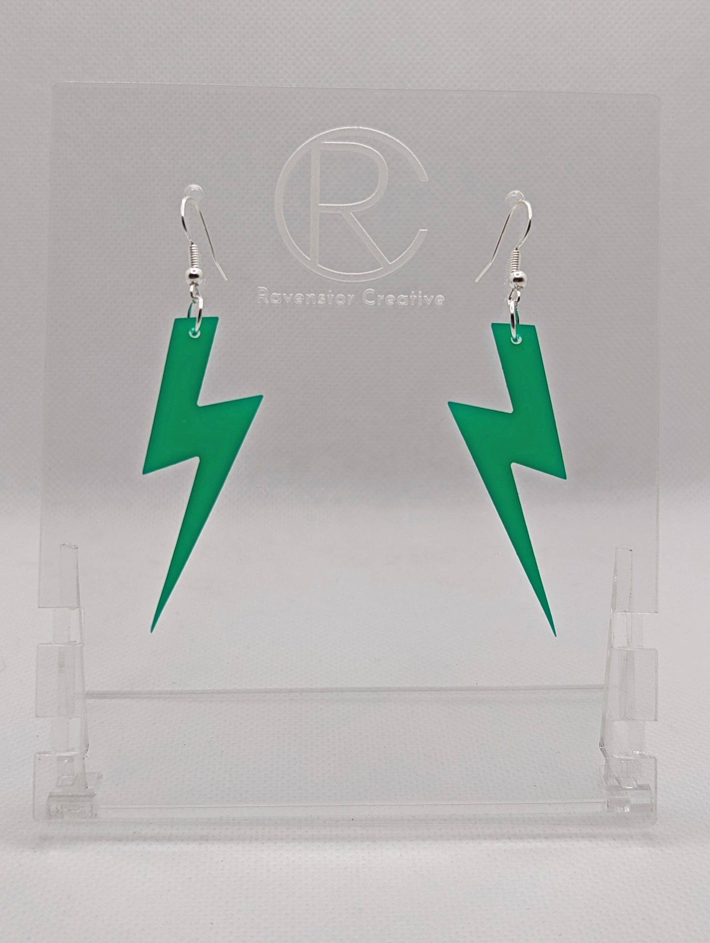 Shows a pair of acrylic earrings that have silver ear wire hook fixings. They are Lightning Bolt shapes similar to Harry Potter's forehead. They are green colour and are against a white background on see through stand.