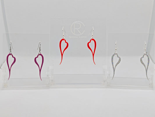 Shows three pairs of earrings that have ear wire hook fixings. They are pretty melting love Heart shaped. The colours are Grape, red and Holographic Glitter. They are against a white background on see through stand.