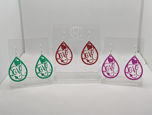 Image of three pairs of teardrop shaped earrings with the word love cut into the middle in a fancy font with a few hearts around it. They have silver fixings and are the colours Green, Red Glitter and Grape on a white background.