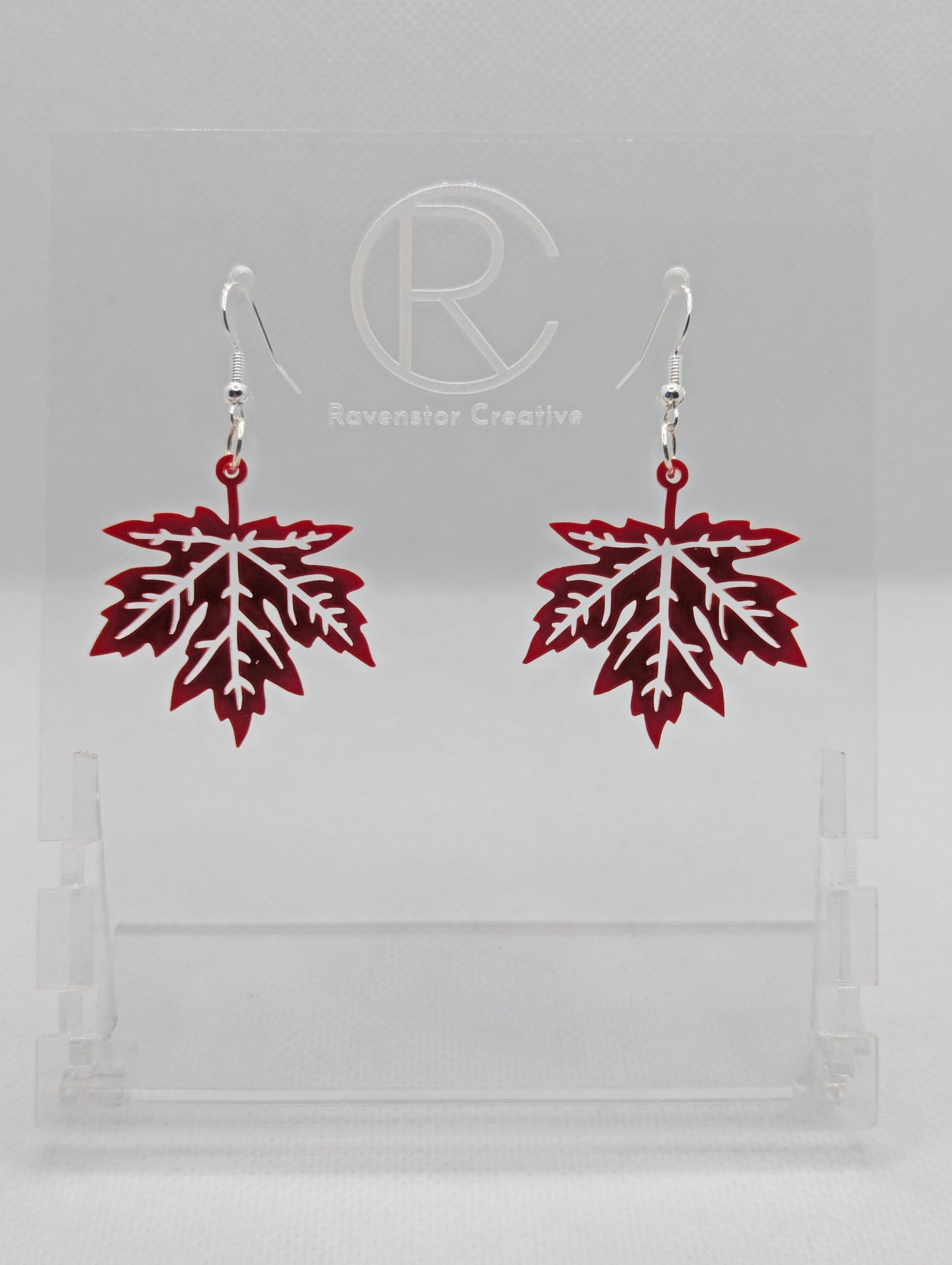 Shows a pair of acrylic earrings that have silver ear wire hook fixings. They are autumn fall maple leaf shapes. They are in Maroon and are against a white background on see through stand with the Ravenstor Creative logo on it.