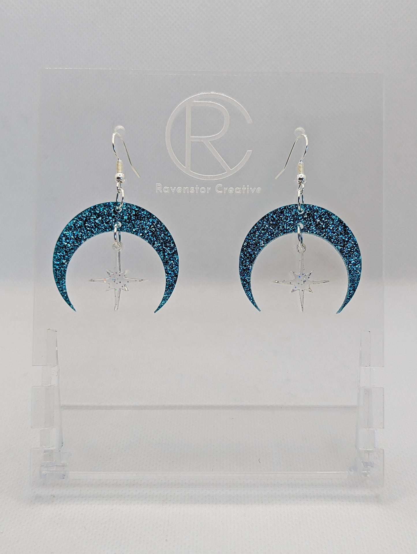 A pair of acrylic earrings that are two parts. Silver ear wire hook fixings are attached to the middle of a sideways crescent moon with a small star dangling in the centre. They have an Ice Blue Glitter Moon with Clear Sparkle star in the middle.