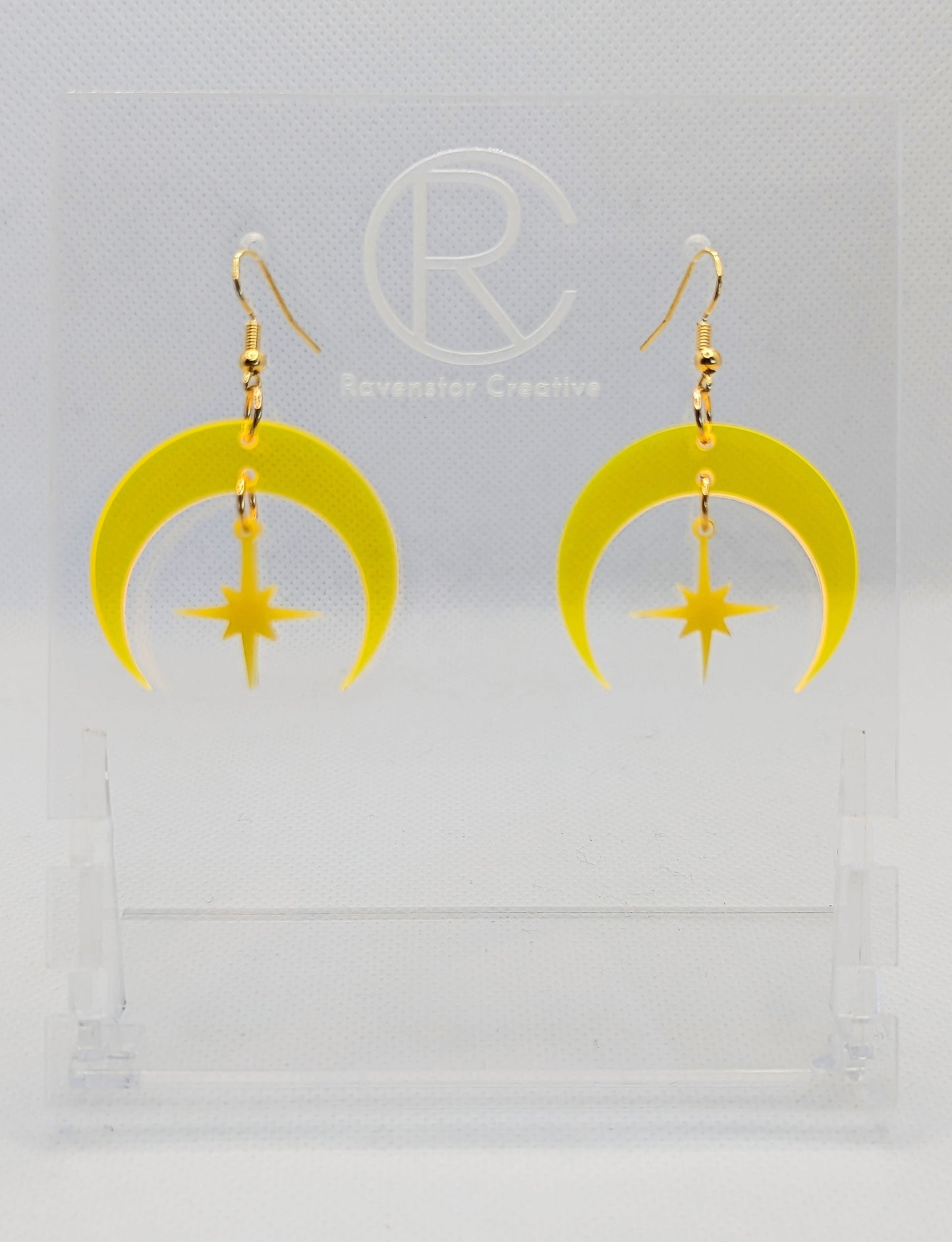 A pair of acrylic earrings that are two parts. Gold ear wire hook fixings are attached to the middle of a sideways crescent moon with a small star dangling in the centre. They have a Neon Orange Moon with a Yellow star in the middle.