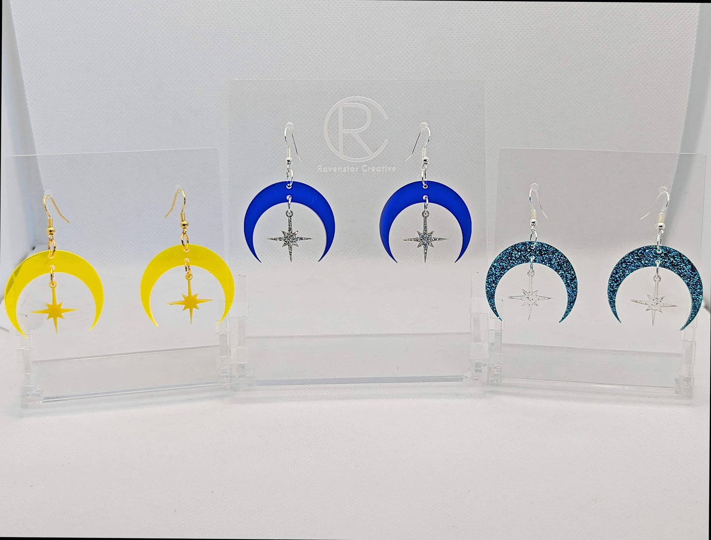 Three pairs of earrings that are two parts. Ear wire fixings are attached to a crescent moon with a small star dangling in the centre. Colours are Neon Orange moon with Yellow star, Blue moon Silver glitter star. Ice Blue moon and Clear sparkle star.